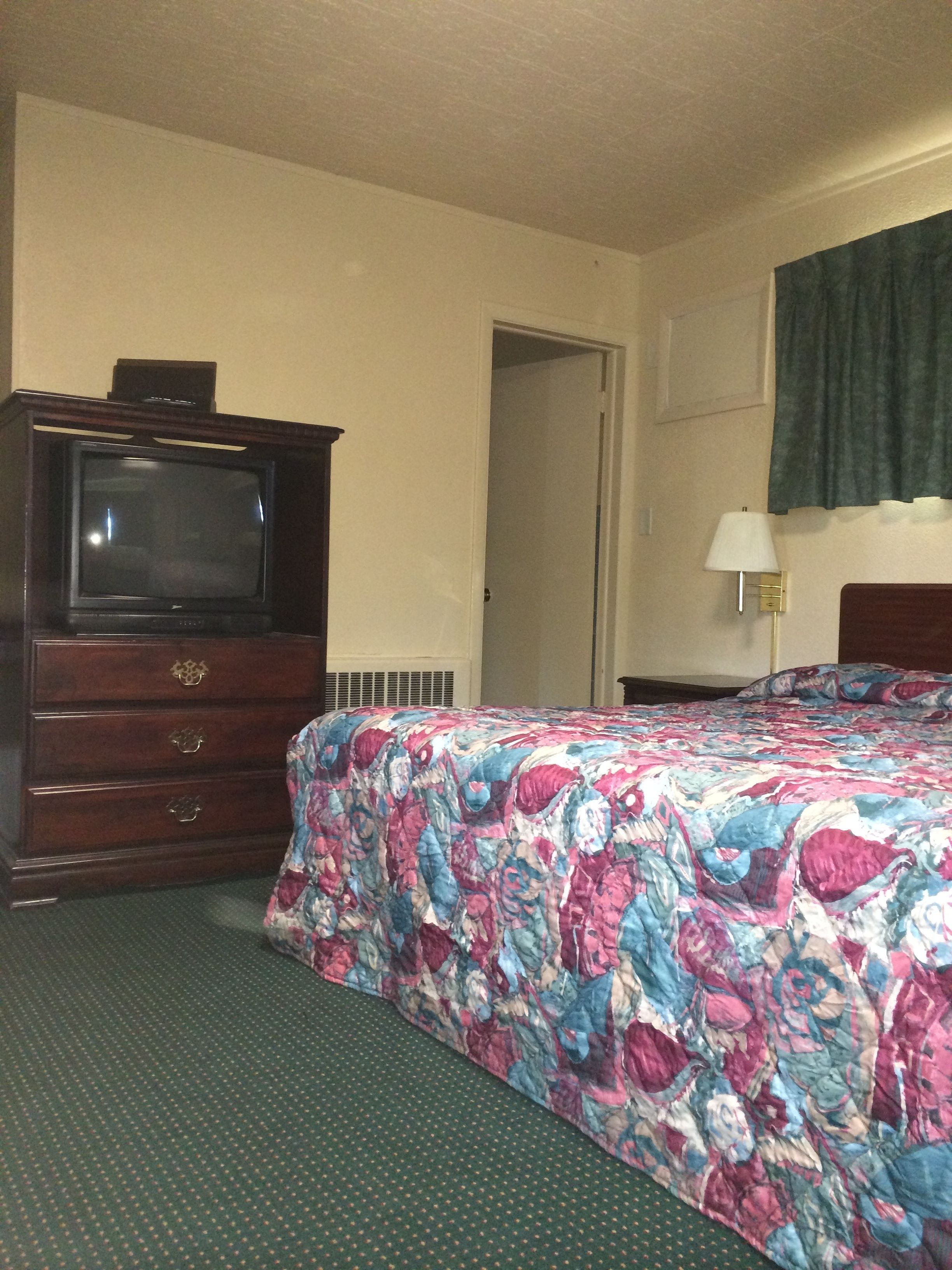 Una habitación de hotel con cama, cómoda, televisor, puerta y lámpara.