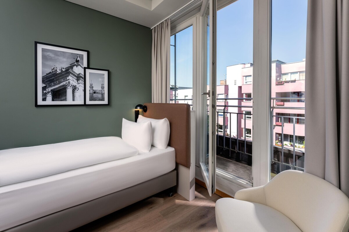 A hotel room with a bed, a chair, a window with a view of buildings, and two framed pictures on the wall.