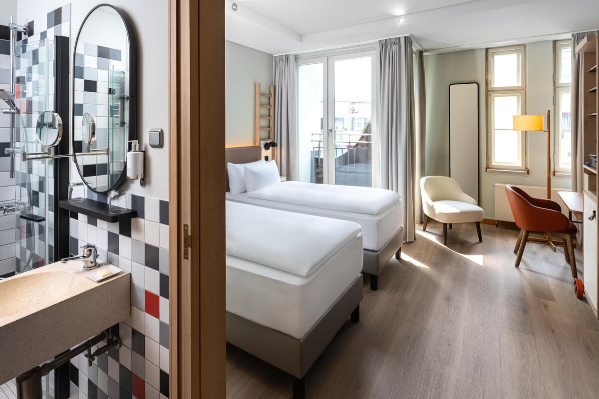 Habitación de hotel con salpicadero de azulejos a cuadros, una cama, una silla, un escritorio y una ventana con cortinas.