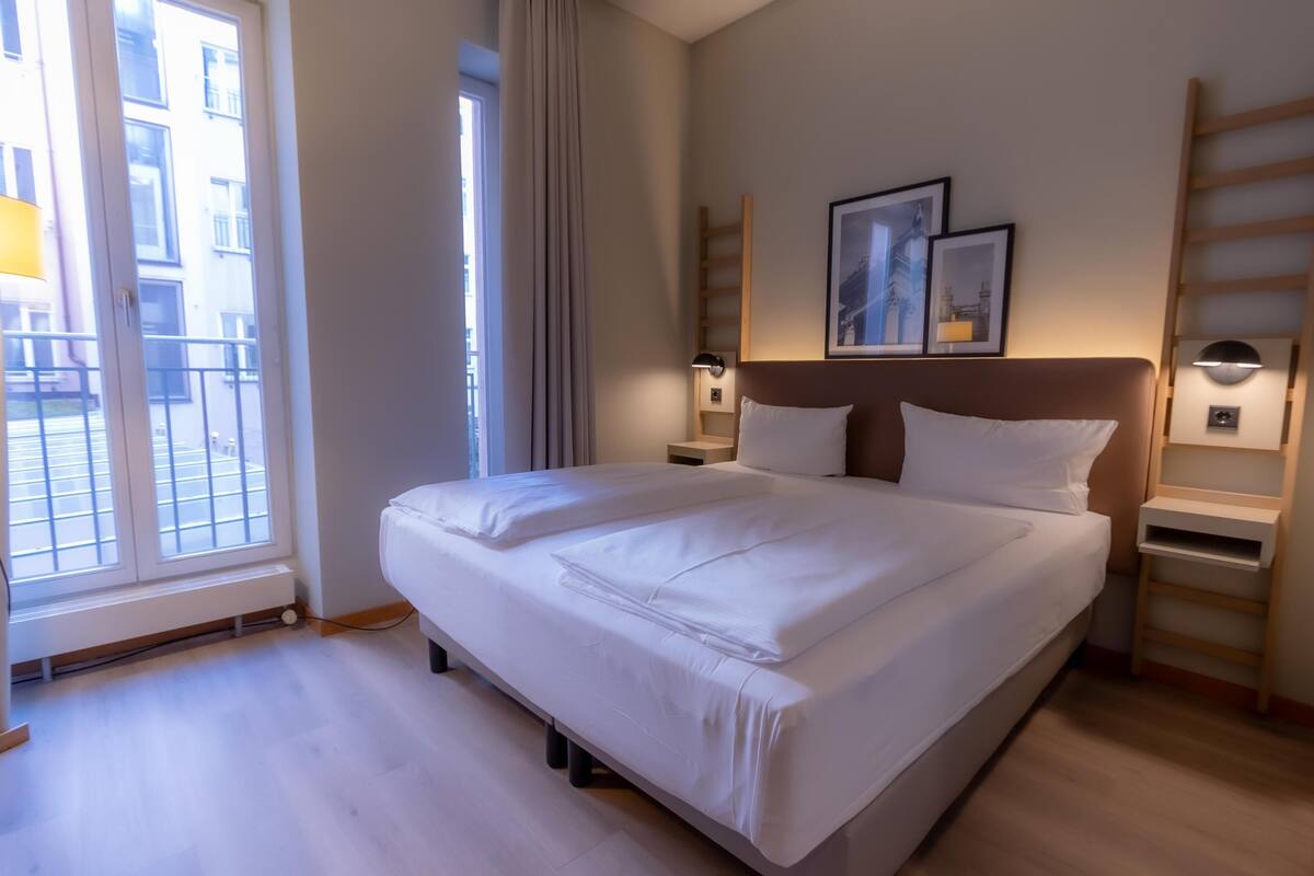 Habitación de hotel con una cama grande, una ventana con cortinas, una mesita de noche con lámpara y un estante en la pared.