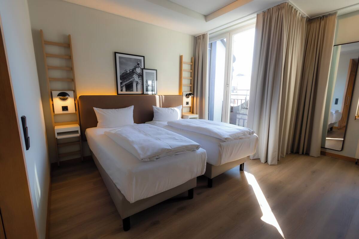 Habitación de hotel con dos camas, una escalera, un cuadro enmarcado, una mesita de noche y vistas al exterior.