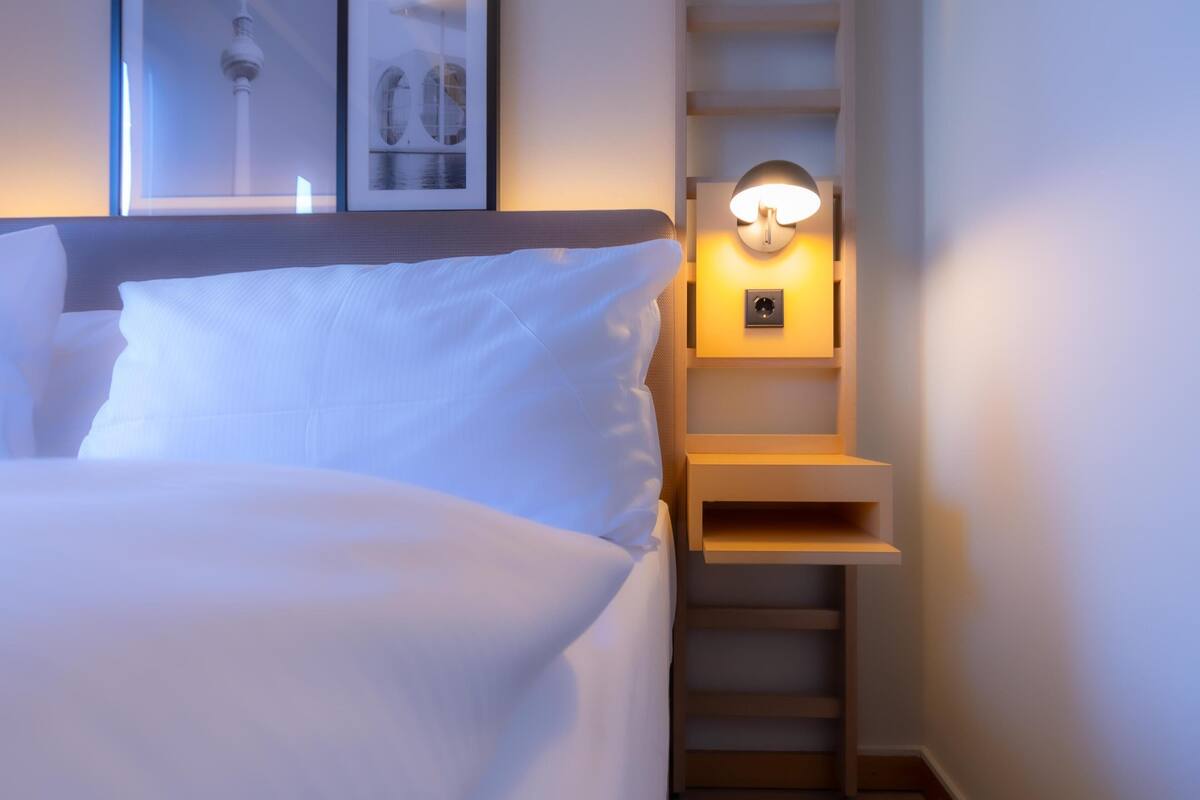 Una cama bien hecha con ropa blanca, un aplique de pared y un cuadro colgado sobre la cama.