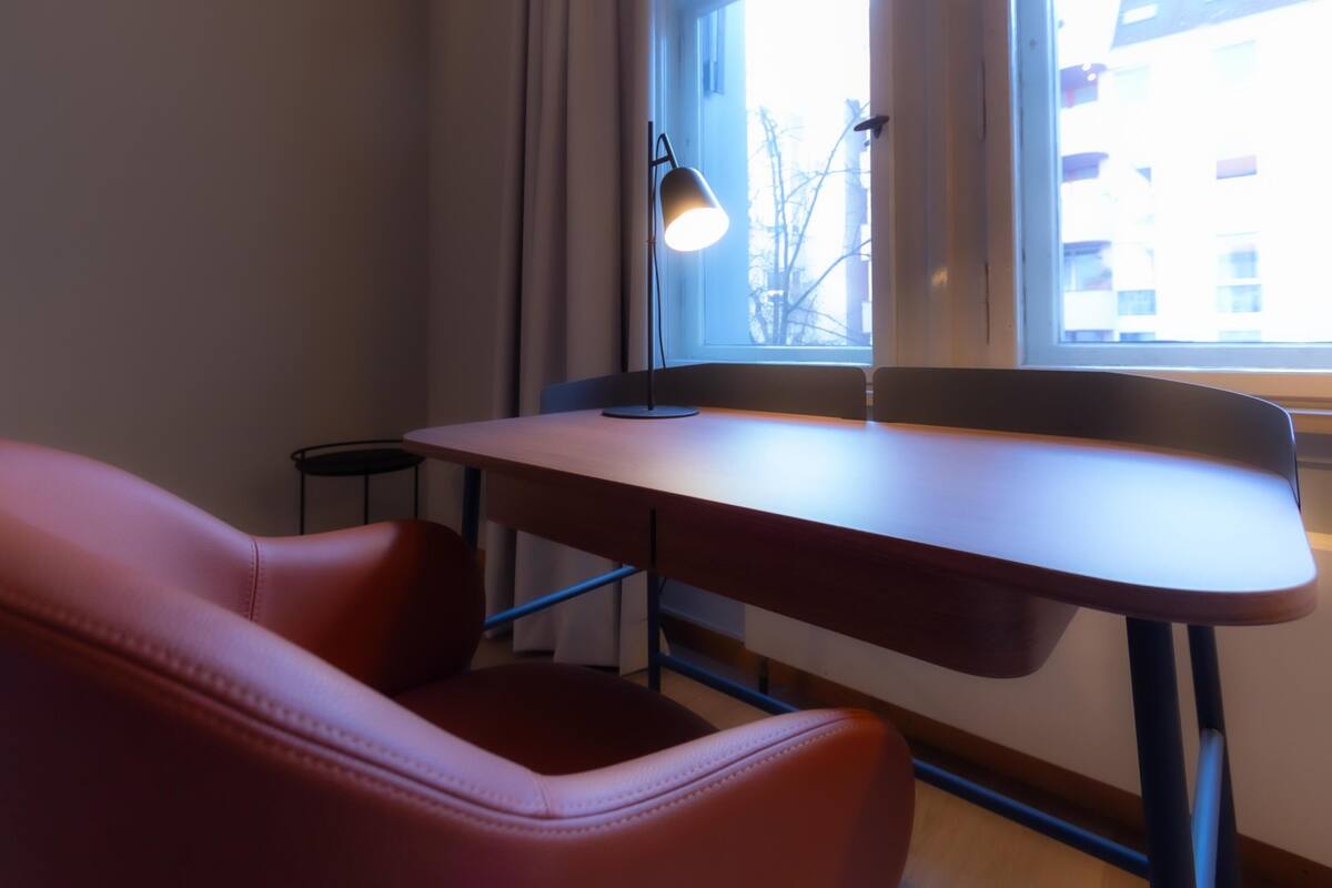 Una habitación con escritorio, silla, lámpara y ventana con vista a edificios.