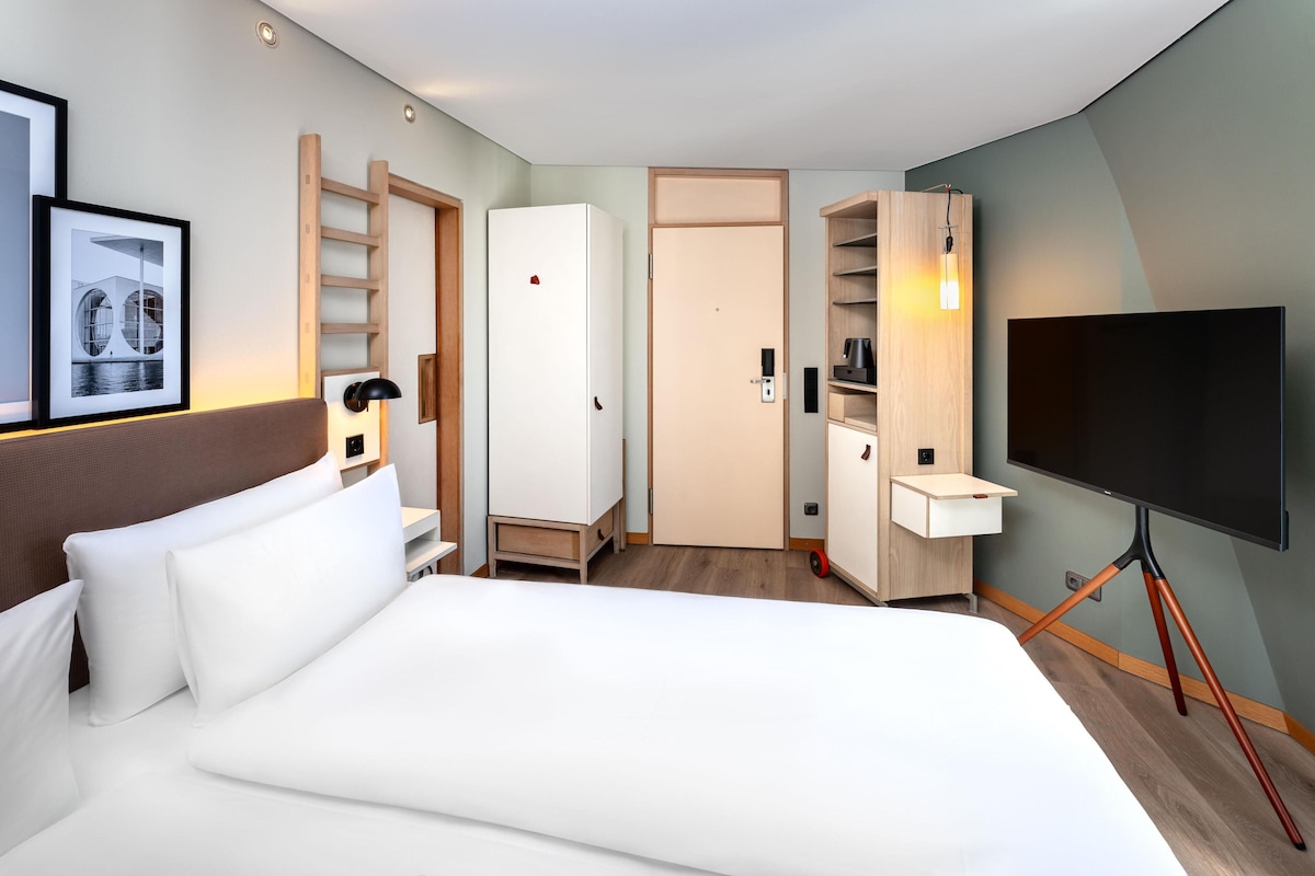 A hotel room with a bed, a TV, a desk, a wardrobe, and a framed picture on the wall.