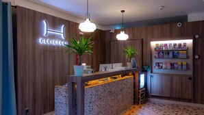 A reception desk with a wooden backdrop, two potted plants, a first aid sign, and a shelf with various items.
