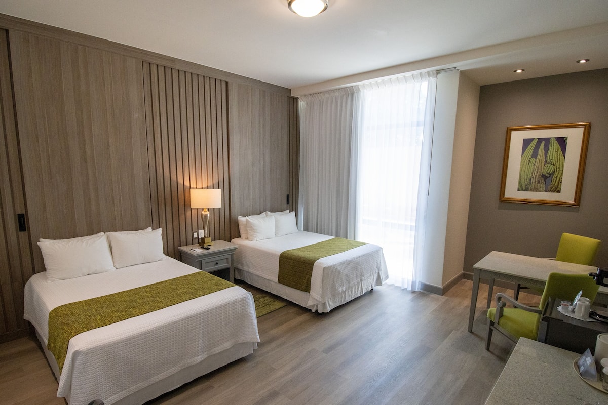 A hotel room with two beds, a desk, a chair, a lamp, and a painting on the wall.