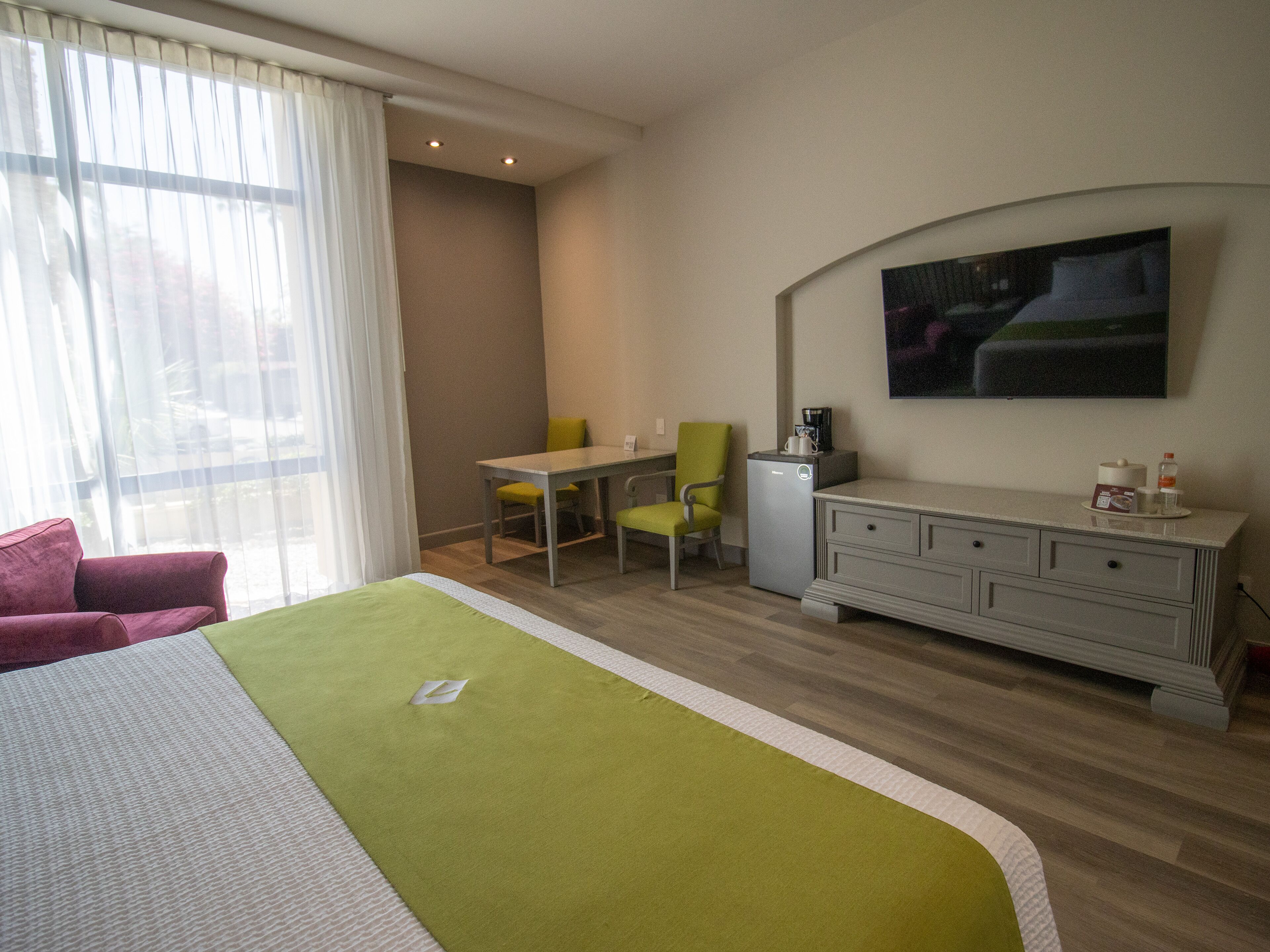 Habitación de hotel con cama, escritorio, silla, refrigerador pequeño y televisión de pantalla plana montada en la pared.