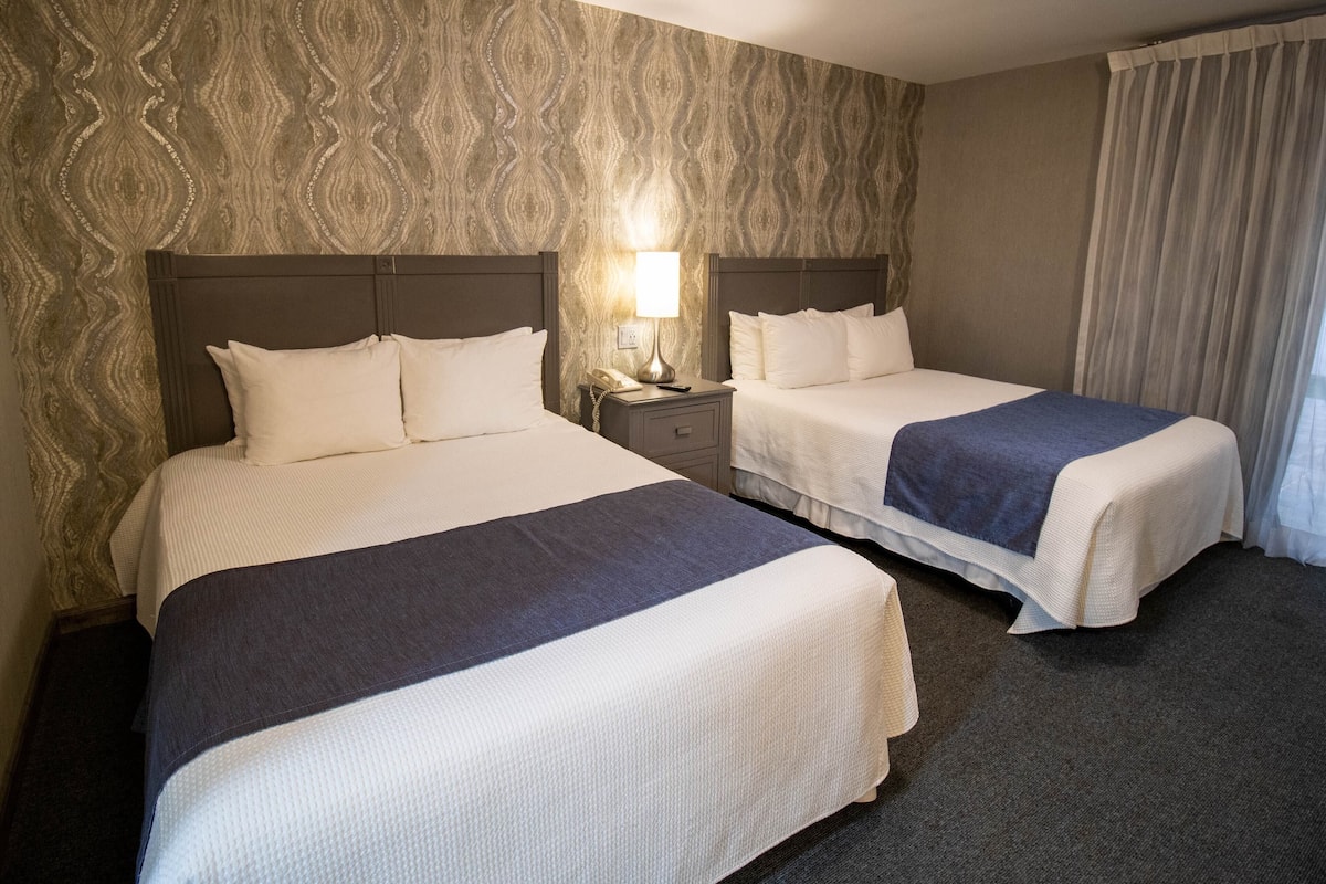 A hotel room with two beds, a nightstand with a lamp, and a patterned wallpaper.