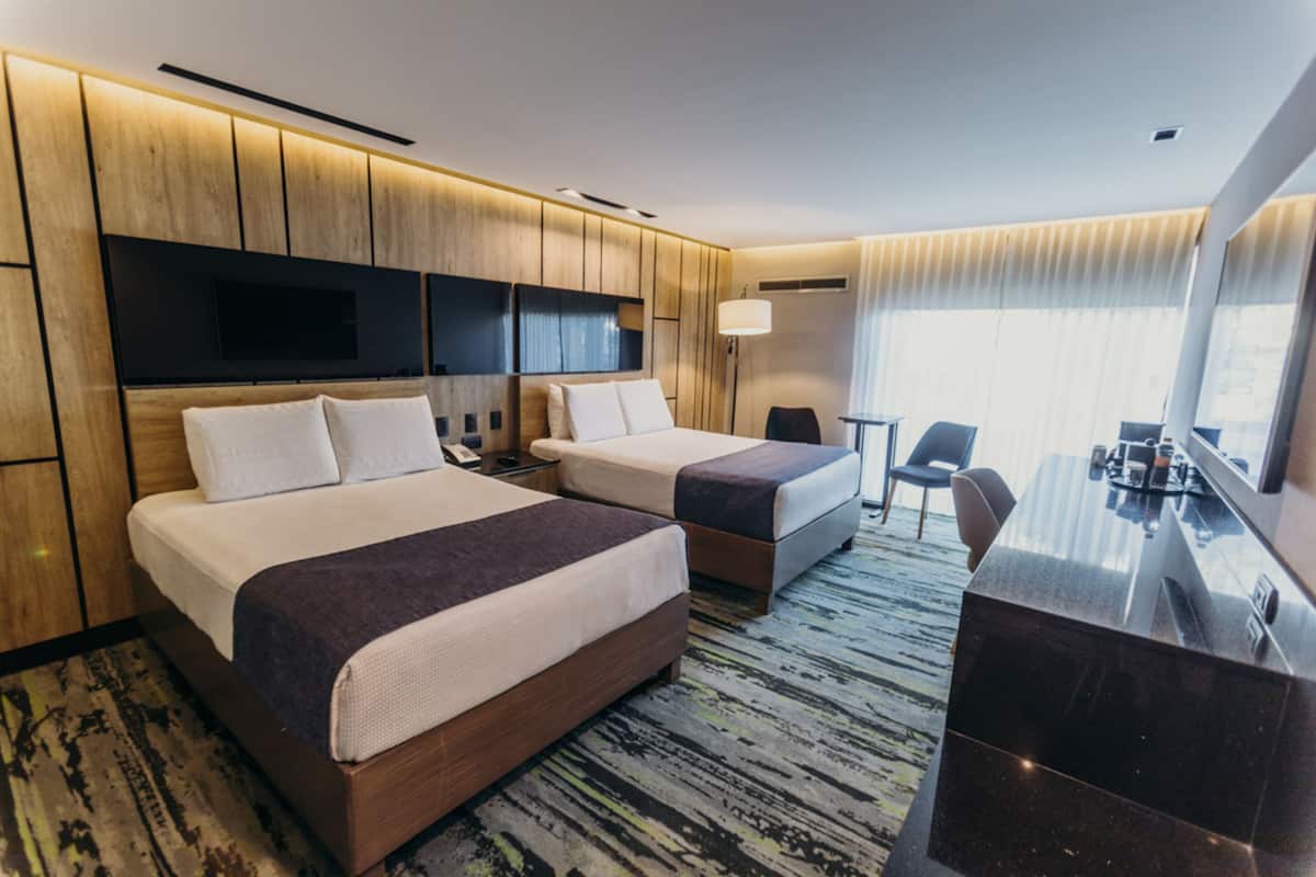 A hotel room with two beds, a desk, a chair, a TV, and a large window with curtains.