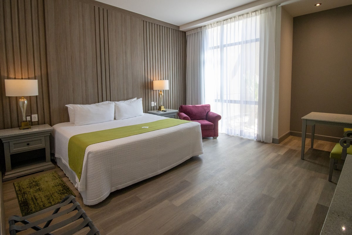 A hotel room with a large bed, two bedside lamps, a chair, a small table, and a window with curtains.