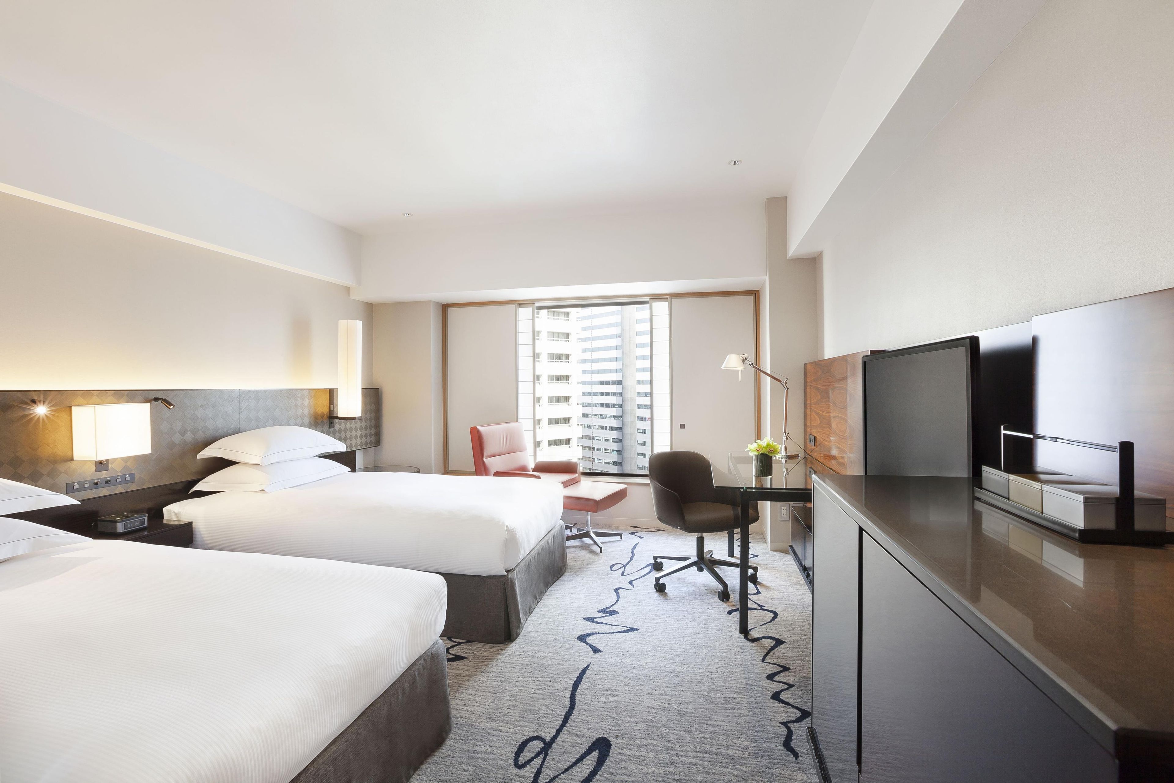 A hotel room with two beds, a desk, a chair, a television, and a large window with a view of buildings.