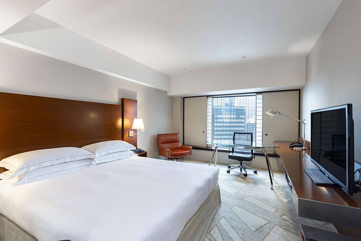 A hotel room with a large bed, a desk, a chair, a TV, and a window with a city view.