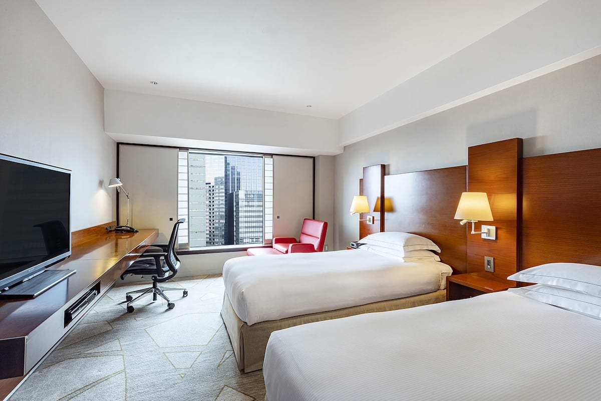 A hotel room with a large flat-screen TV, two beds, a desk with a computer, a red chair, and a window with a city view.
