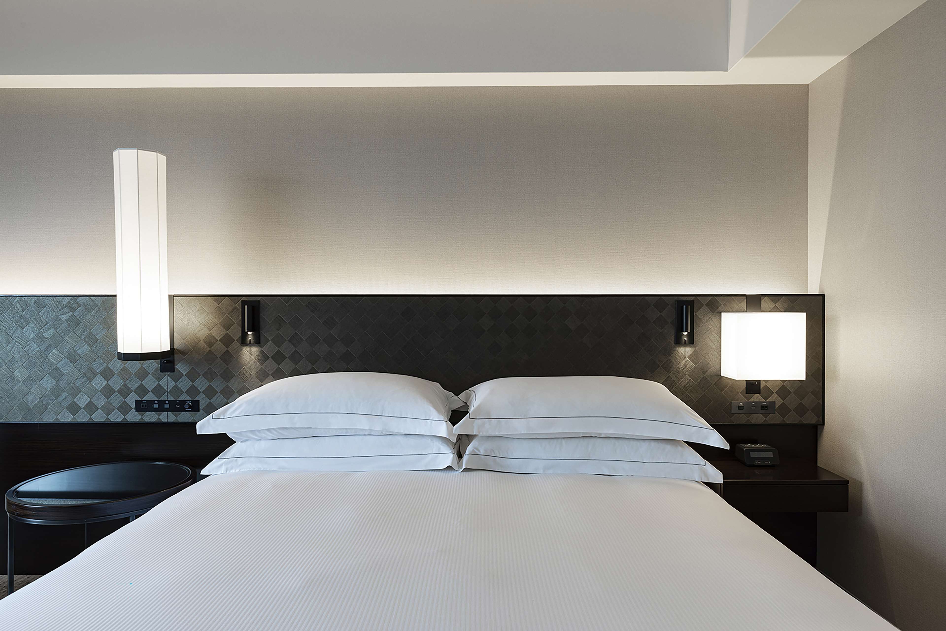 A hotel room with a large bed, two bedside lamps, a small table, and a wall-mounted control panel.
