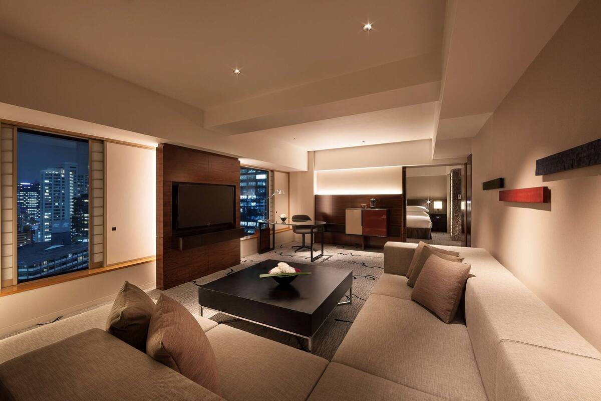 A modern hotel room with a large TV, a seating area with a sofa and coffee table, a desk, and a bed.