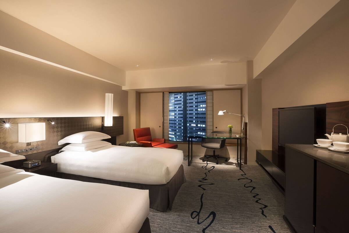 A hotel room with two beds, a desk, a TV, and a view of a cityscape through the window.