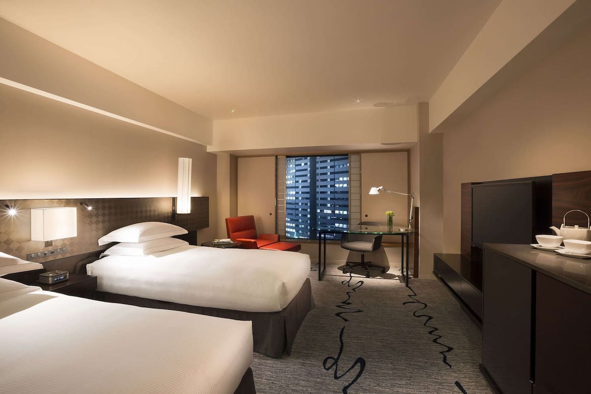 A hotel room with two beds, a desk, a TV, and a view of a cityscape through the window.