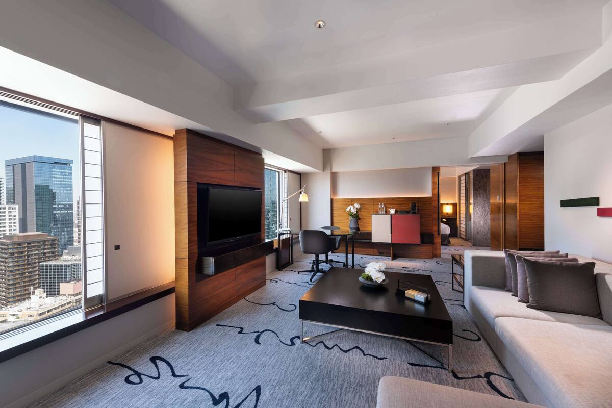 A modern hotel room with a large TV, a sofa, a coffee table, and a view of the cityscape.