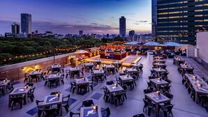 An outdoor dining area with tables and chairs, a bar, and a city skyline in the background. - Hilton Tokyo (Tokyo)