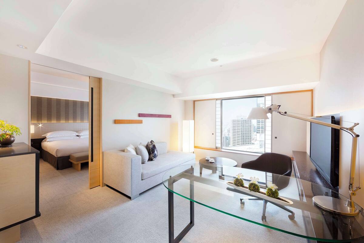 A modern hotel room with a bed, a sofa, a glass coffee table, a desk, and a TV.