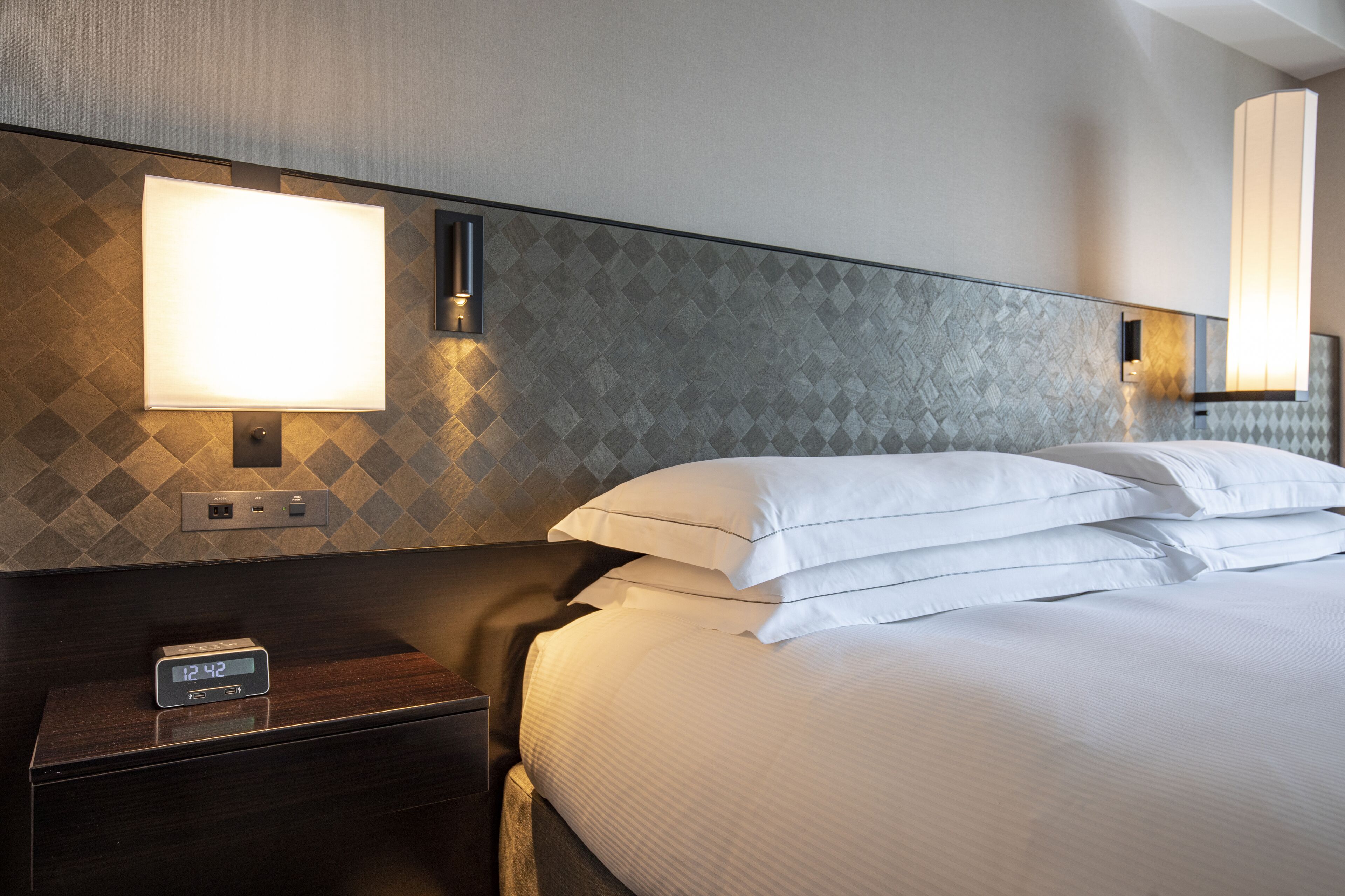 A hotel room with a bed, a nightstand with a digital clock, and a wall-mounted light fixture.