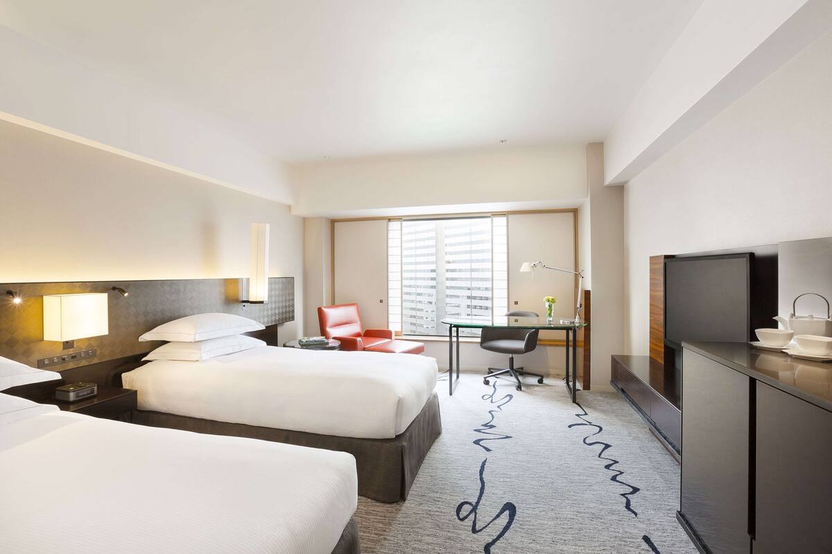 A hotel room with two beds, a desk, a chair, a TV, and a window with blinds.