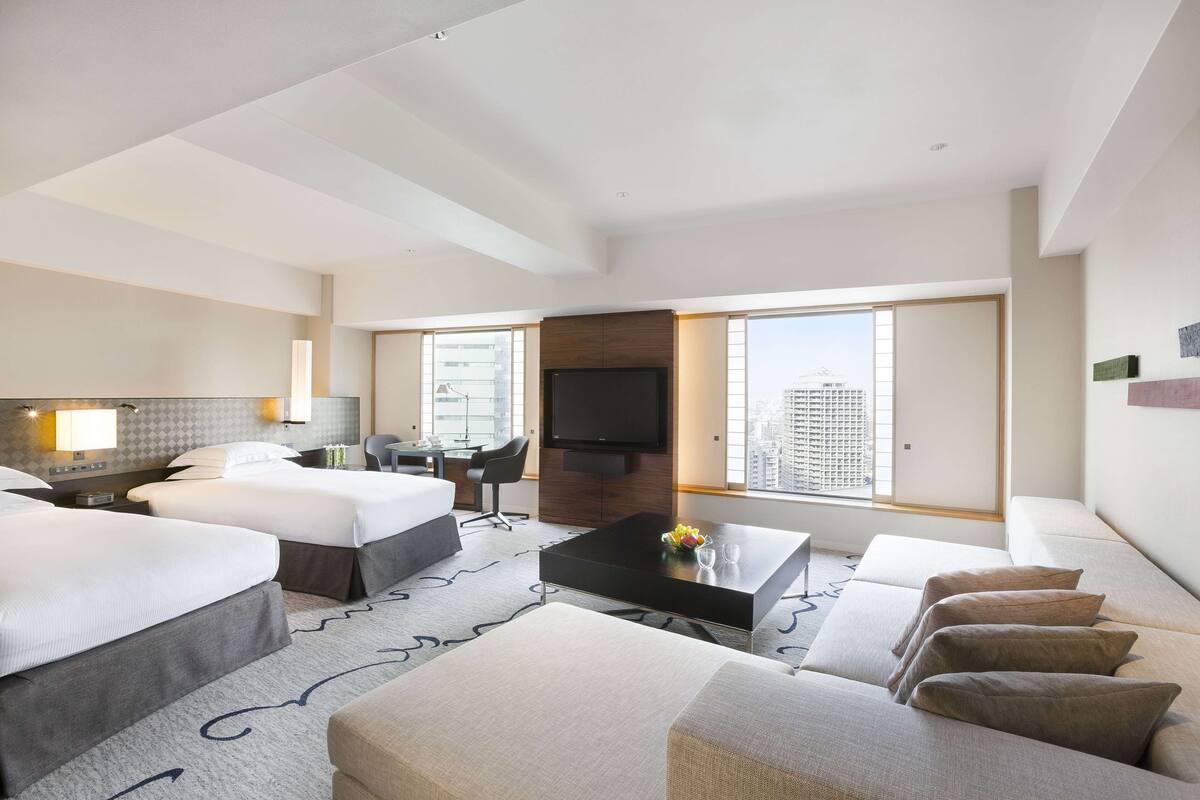A hotel room with two beds, a flat-screen TV, a desk, and a large window offering a city view.