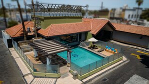 A rooftop swimming pool with a glass enclosure, a covered seating area, and a building with a red-tiled roof.