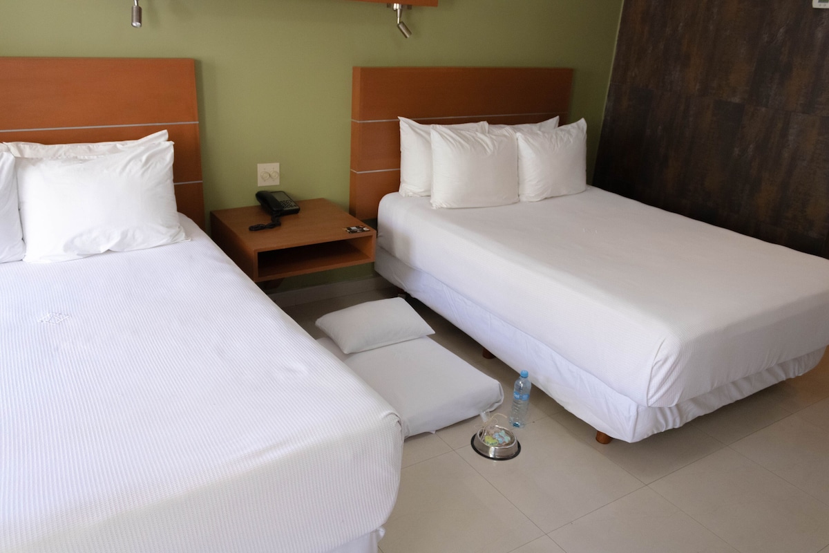 A hotel room with two single beds, a wooden nightstand, a small table with a phone and remote control, and a water bottle on the floor.