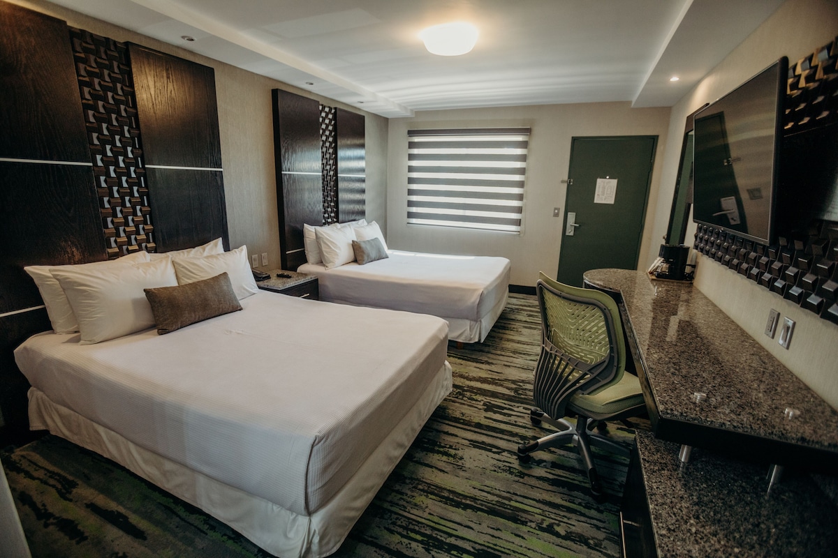 A hotel room with two beds, a desk, a chair, a television, and a window with blinds.