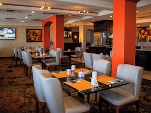 A hotel dining area with tables and chairs, a bar, and a television screen.