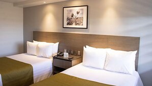 A hotel room with two beds, each with white linens and pillows, a wooden headboard, a nightstand with a phone and a small table, and a framed picture on the wall.