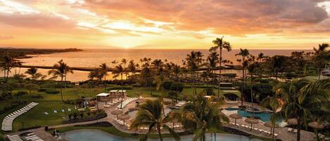 Un resort con una piscina grande, palmeras y vista al océano al atardecer.