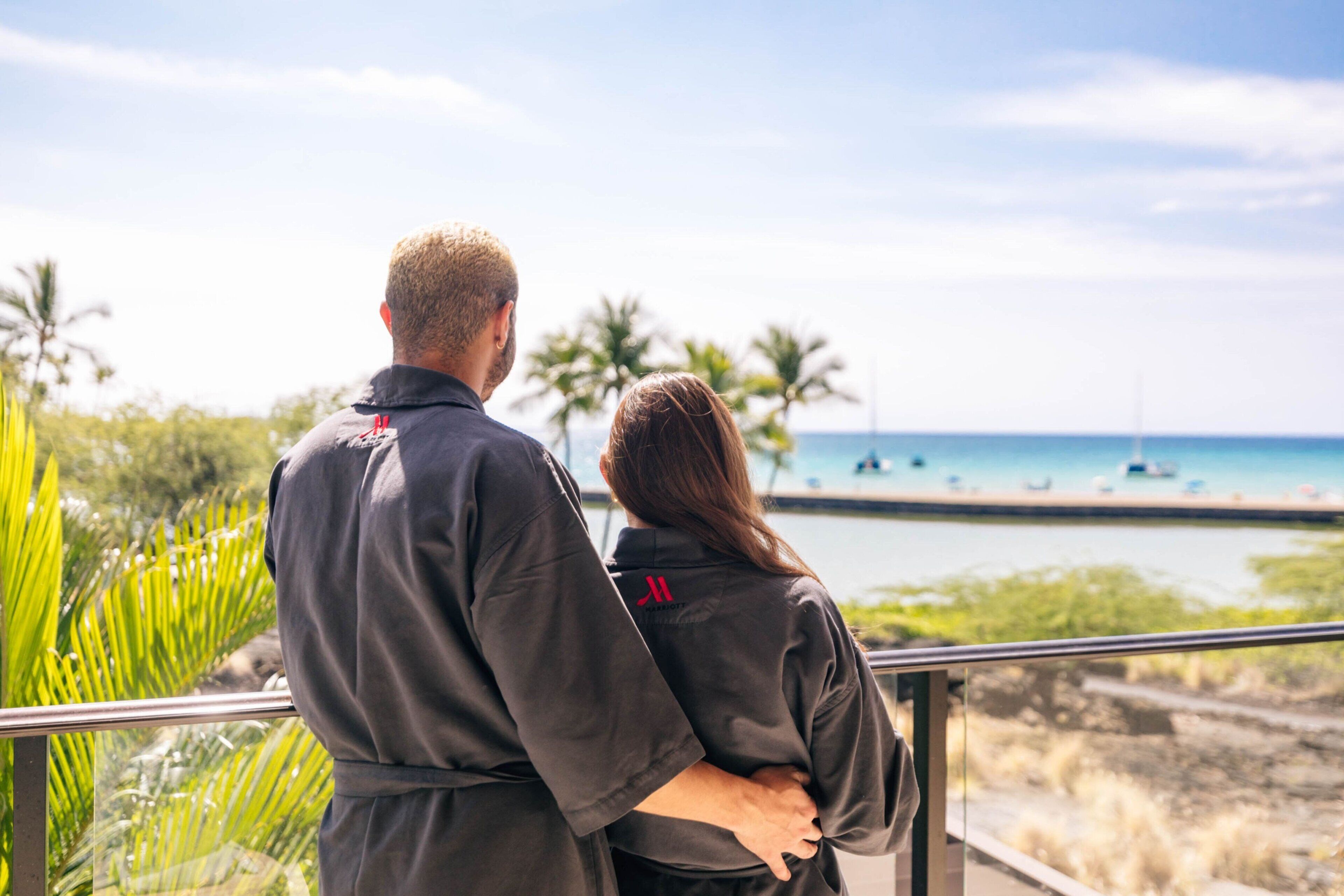 A couple standing on a balcony overlooking a beach with palm trees and boats.