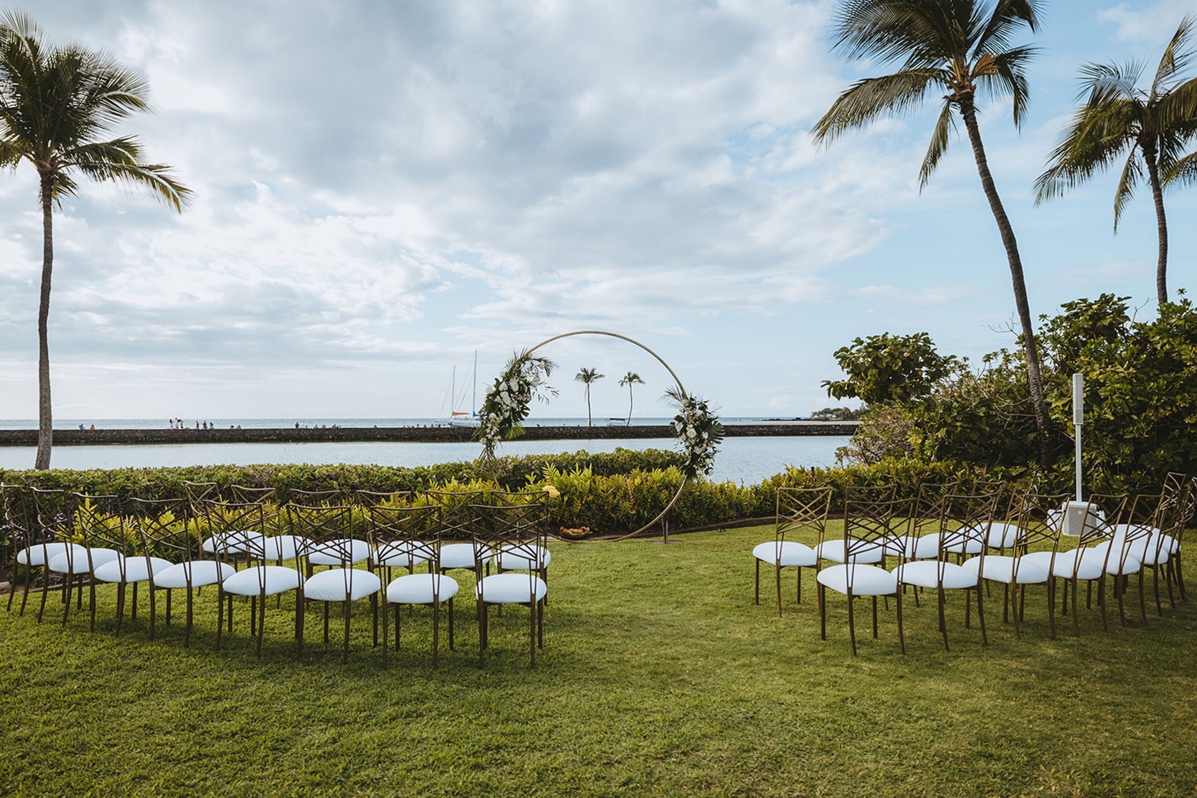 An outdoor wedding setup with chairs arranged in rows, a floral arch, and palm trees.