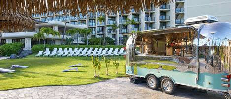 A food truck with a transparent canopy parked in front of a hotel with a pool and lounge chairs.