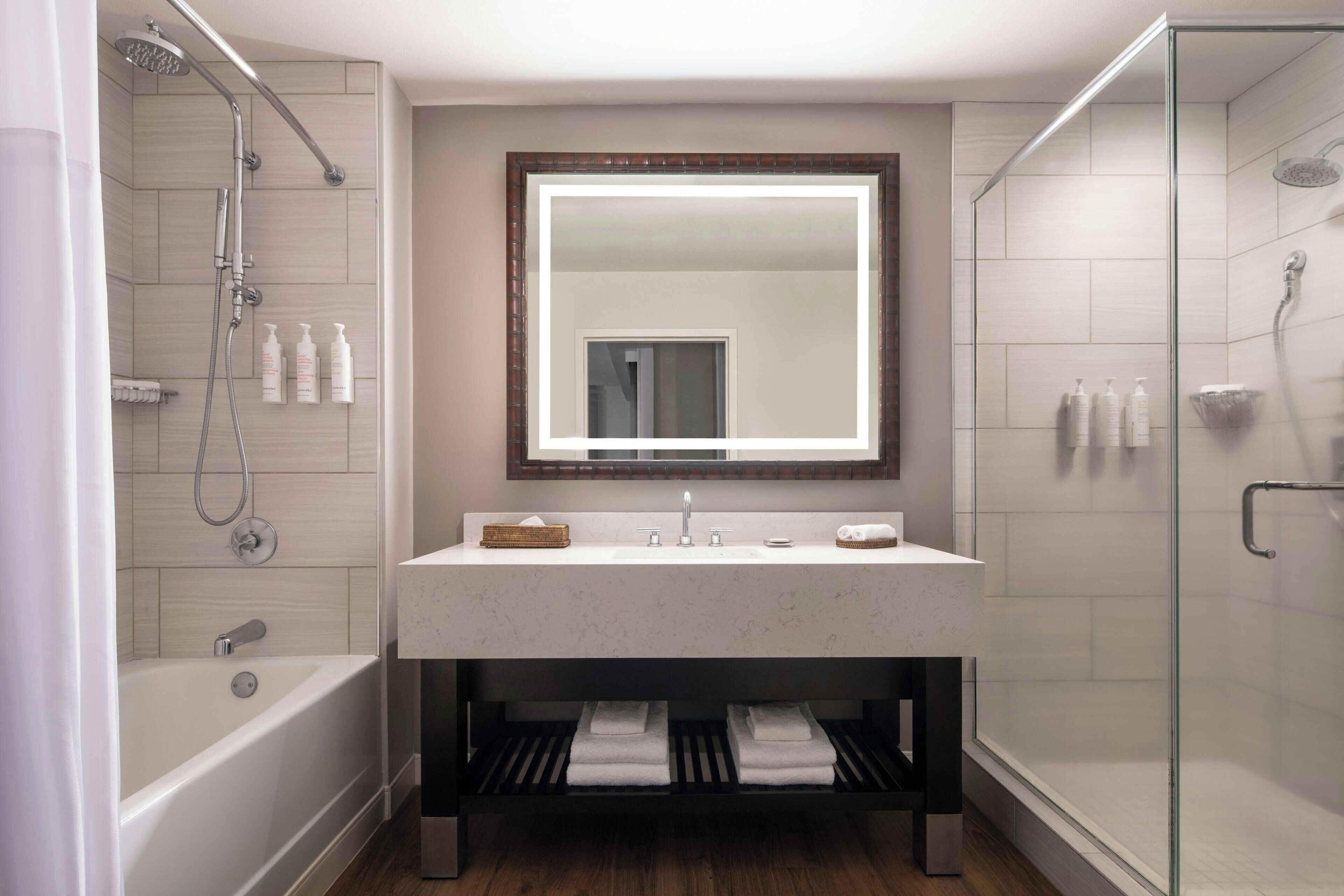 A modern bathroom with a glass-enclosed shower, a large mirror above the sink, and a bathtub with a wooden shelf below.