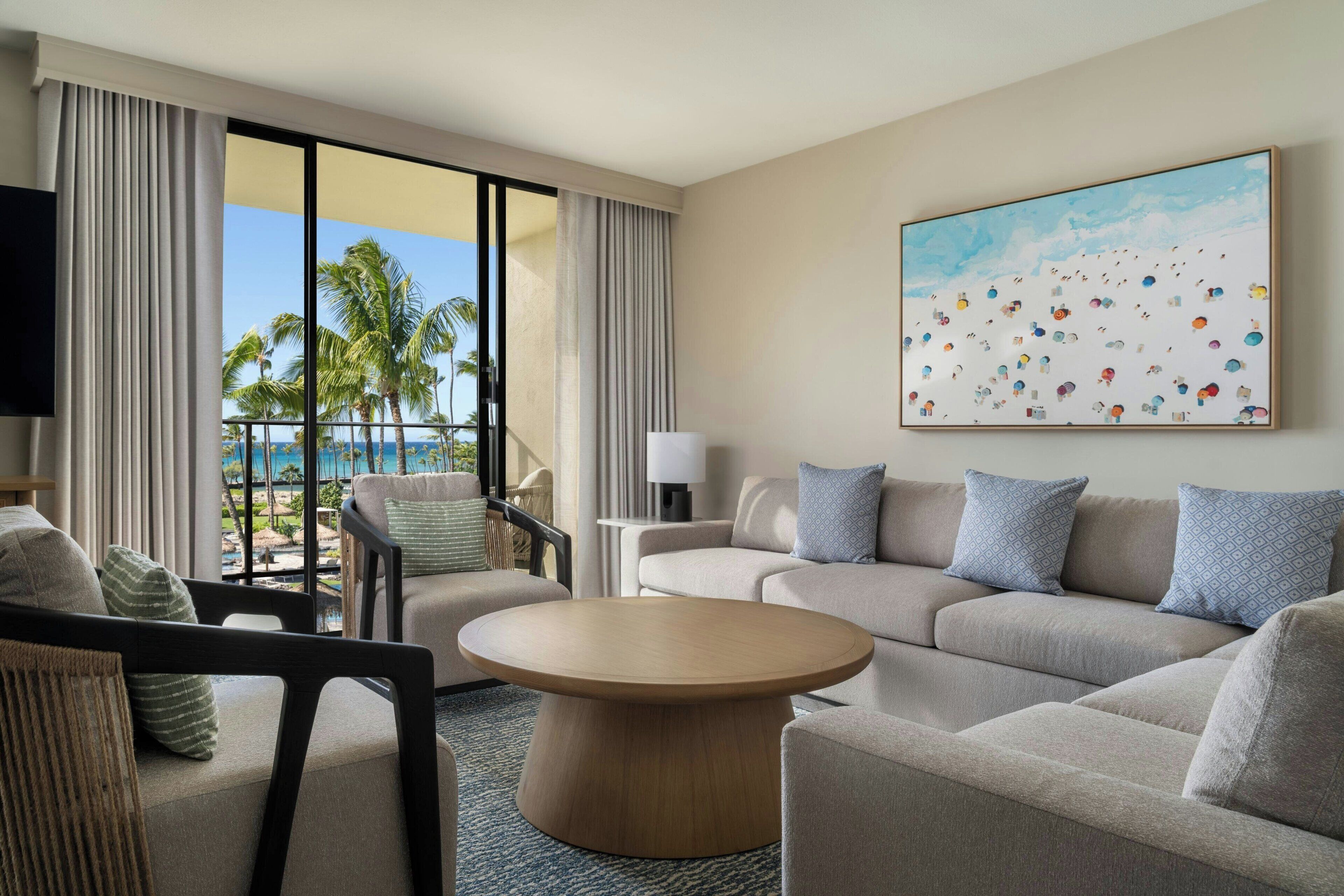 Une chambre d’hôtel dotée d’une grande fenêtre, d’un ensemble de canapés, d’une table basse ronde et d’un tableau représentant une scène de plage.