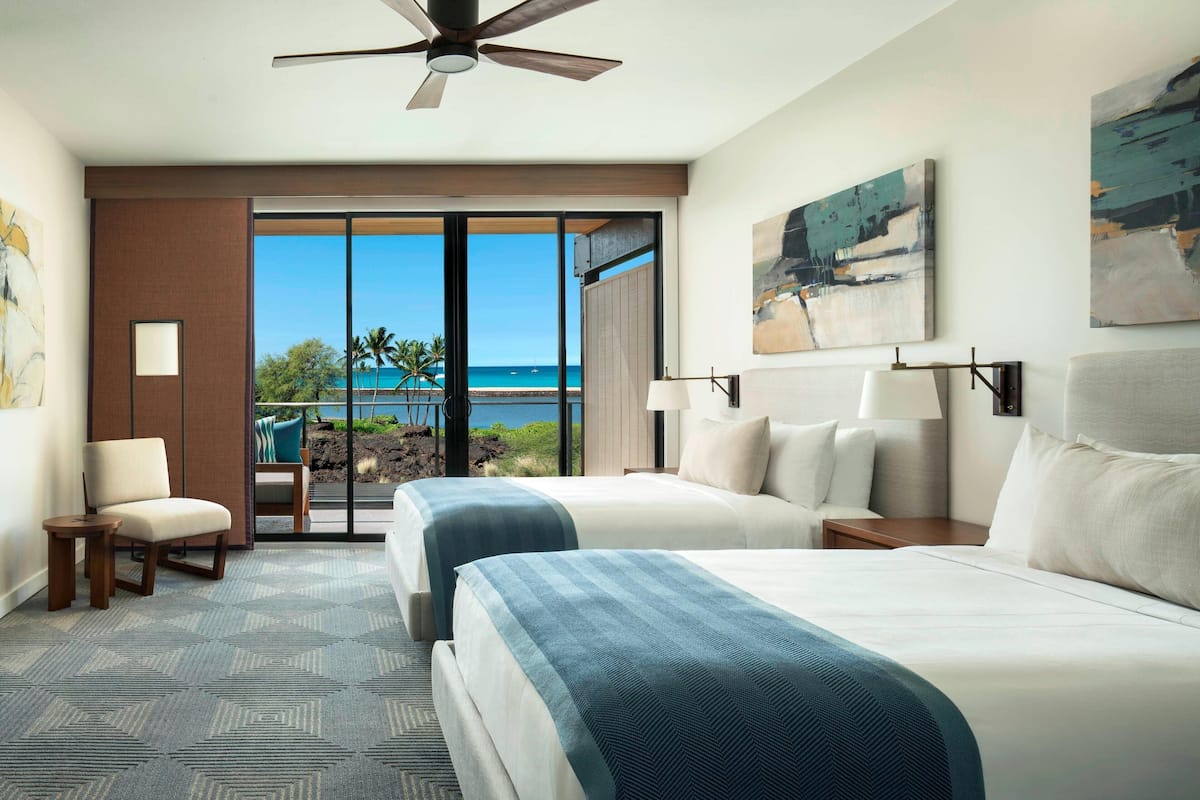 A hotel room with two beds, a ceiling fan, a large window with a view of the beach, and a painting on the wall.