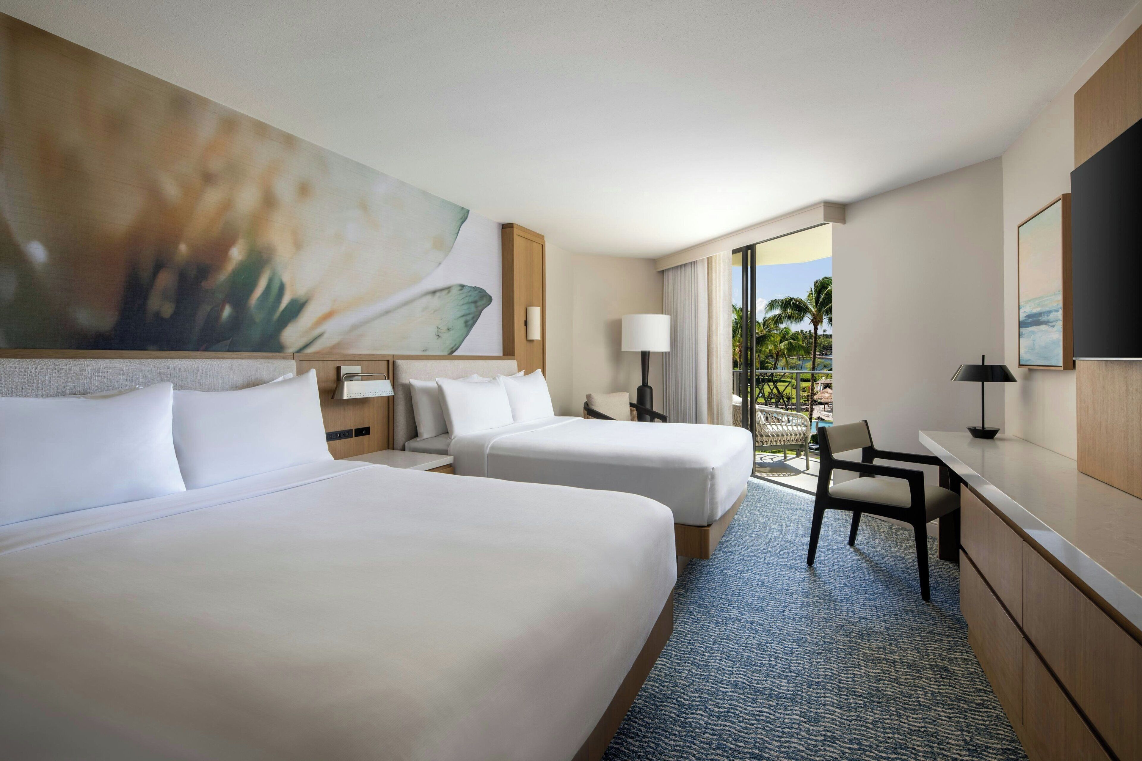 A hotel room with two beds, a desk, a chair, and a view of palm trees.