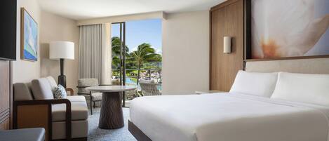 A hotel room with a large bed, a sofa, a small table, and a view of a pool and palm trees.