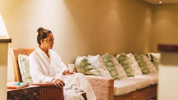 A person sitting on a wicker sofa with a white bathrobe, holding a cup, in a room with a bed and pillows in the background.
