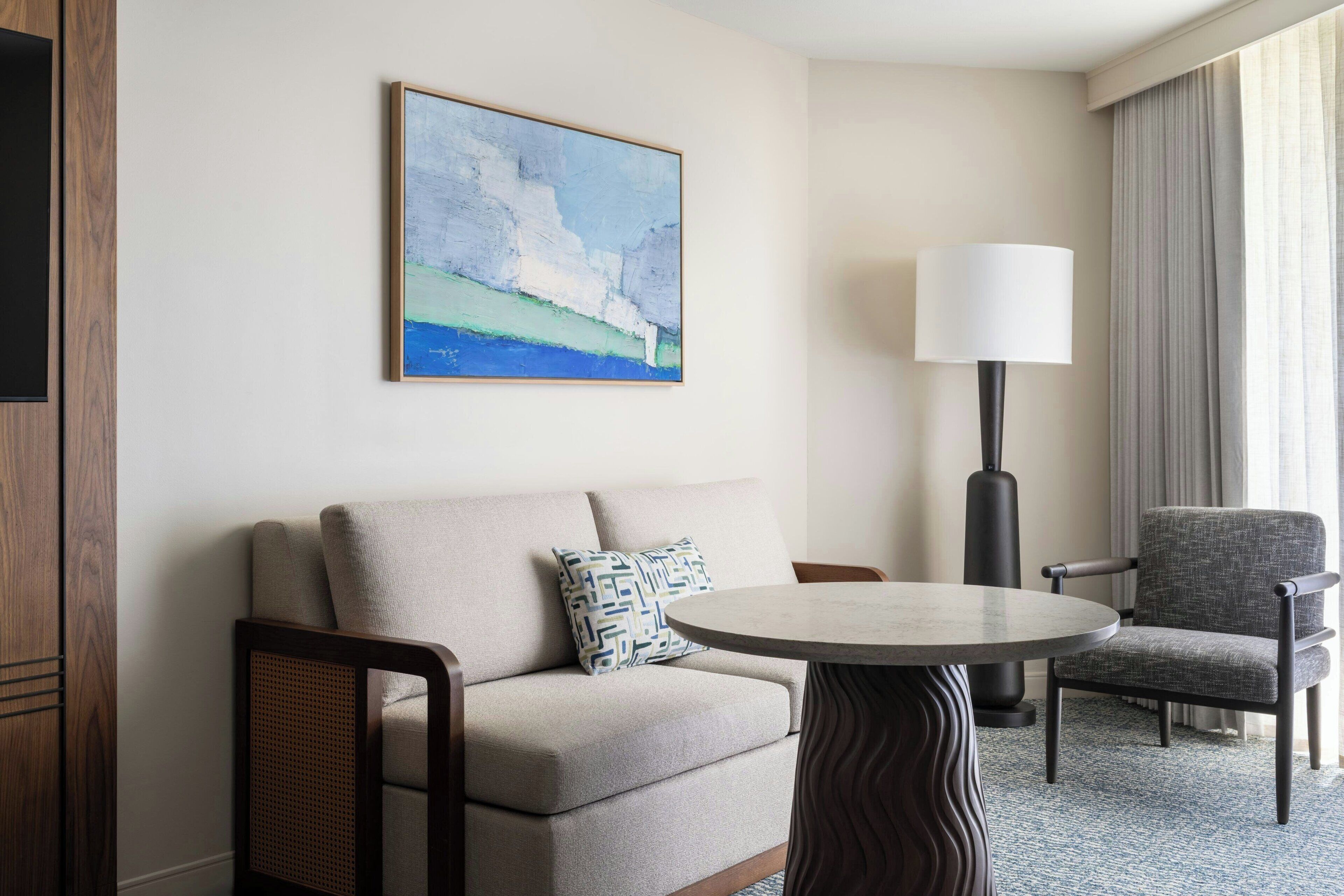 A hotel room with a sofa, armchair, round table, and a painting on the wall.