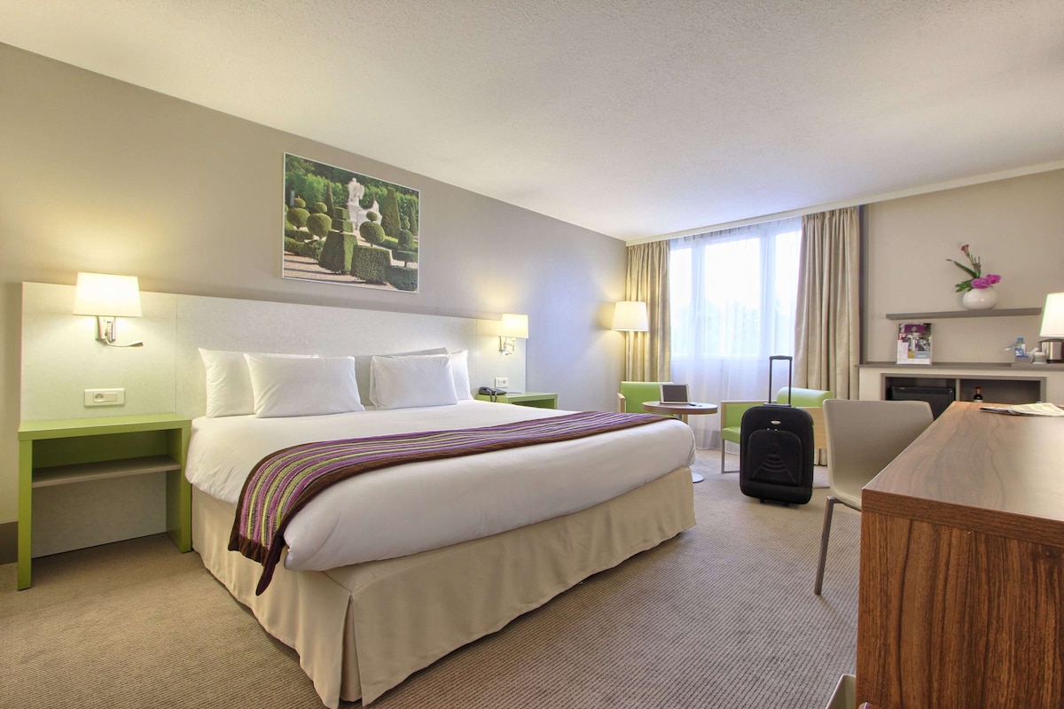 Une chambre d’hôtel avec un grand lit, deux tables de chevet avec des lampes, un bureau avec une chaise, une valise et une fenêtre avec des rideaux.