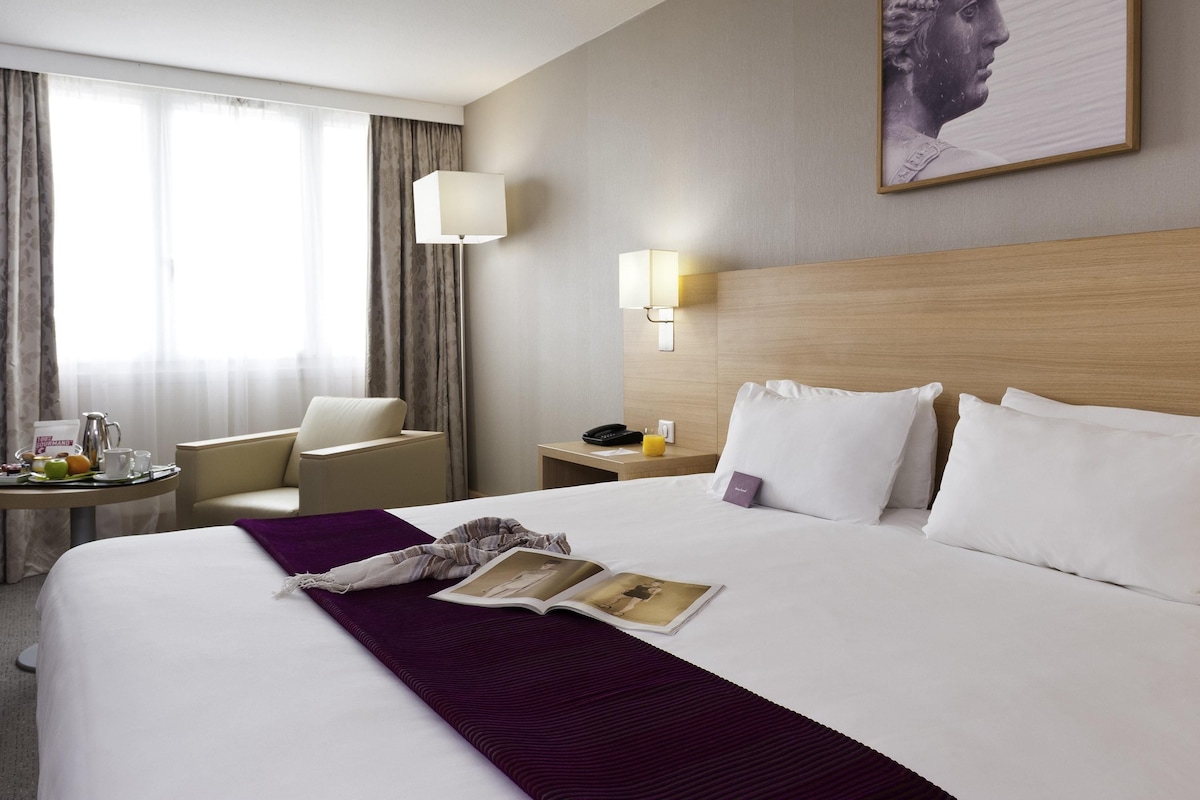 Een hotelkamer met een groot bed, een nachtkastje met een lamp, een stoel en een raam met gordijnen.