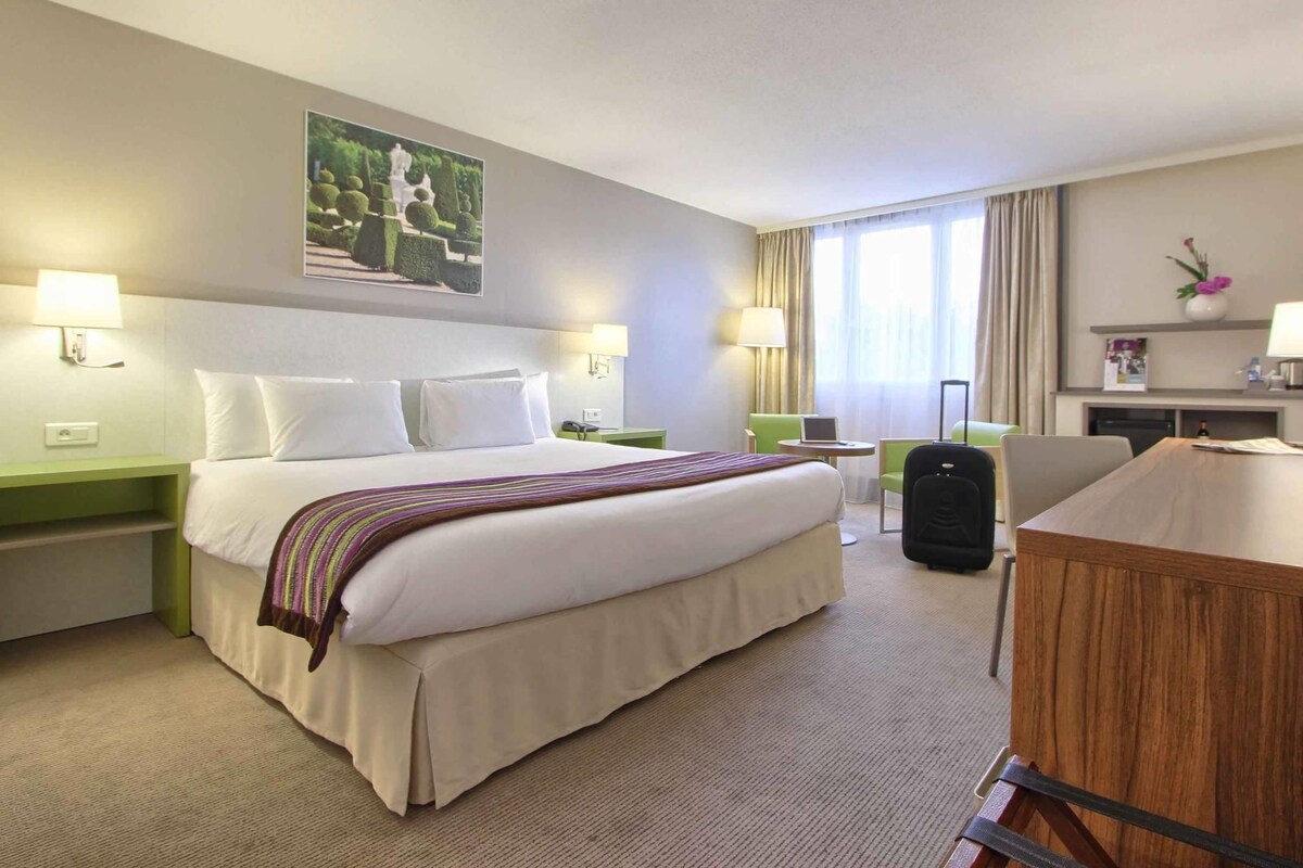 A hotel room with a large bed, two bedside tables, a desk, a chair, a lamp, a picture on the wall, and a window with curtains.
