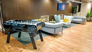Un salon moderne avec une table de baby-foot, un canapé gris et un téléviseur fixé au mur.