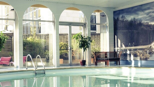 A swimming pool with clear water, surrounded by arched windows and potted plants. - Mercure Paris Velizy (Velizy-Villacoublay)