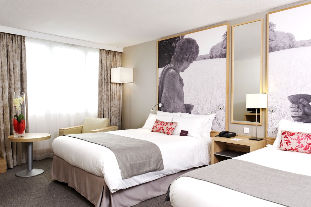 Une chambre d’hôtel avec deux lits, un bureau, une chaise, une petite table avec un vase et une grande fenêtre avec des rideaux.