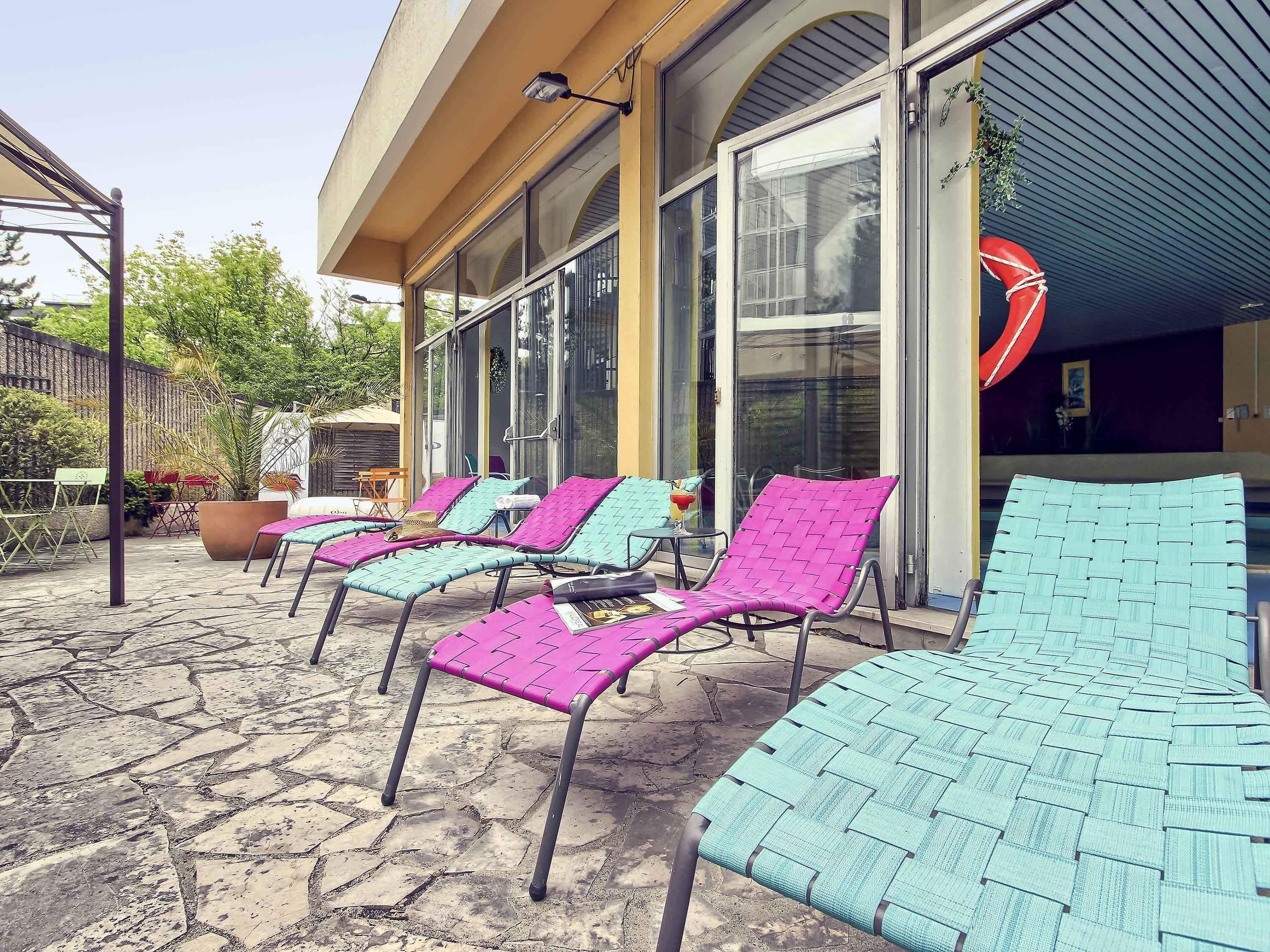 Des fauteuils de jardin avec une structure en métal et des assises en fibres tressées, une terrasse en pierre et un bâtiment doté de grandes fenêtres et d’une entrée abritée.